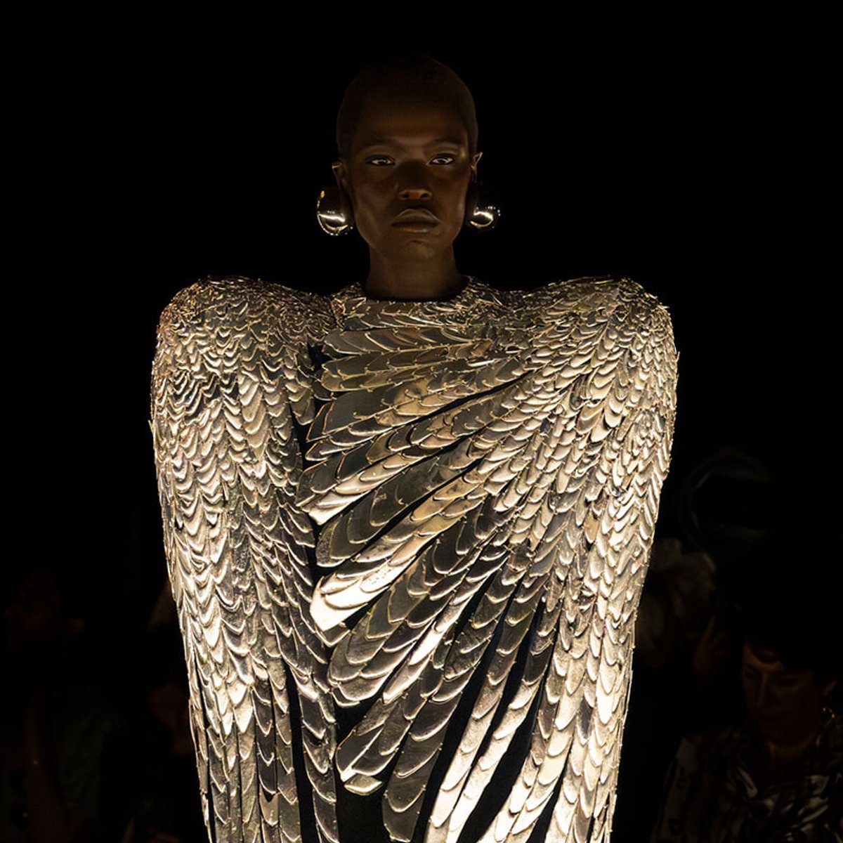 Model wearing a dramatic metallic feathered gown that glow against a dark background on a runway independent of other people visible in the frame.