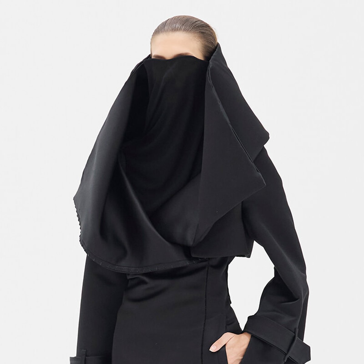 Person wearing a black coat with a large, draped scarf or shawl wrapped over the head and shoulders, obscuring the face against a light background.