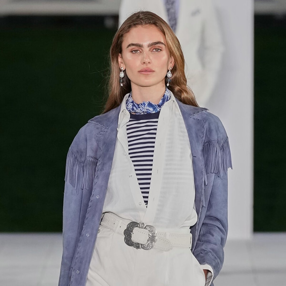 Model on a runway wearing a blue fringe suede jacket over a layered white outfit, with a striped shirt and blue scarf at the neck, accessorized with a white belt and silver buckle.