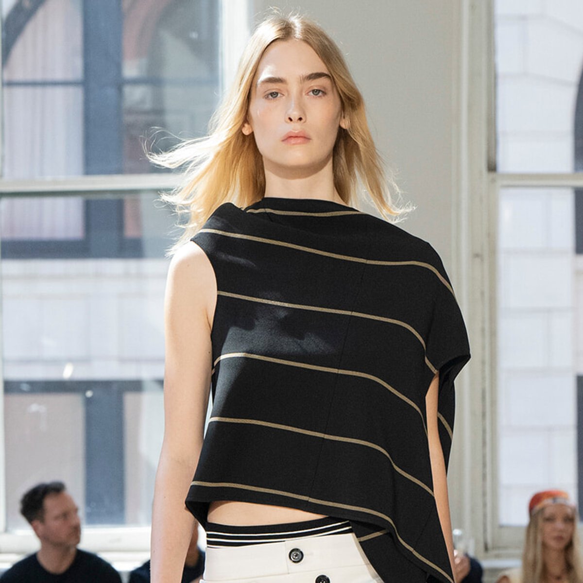Model wearing a black asymmetrical top with beige stripes and white high-waisted pants with buttons, walking the runway in a bright studio.