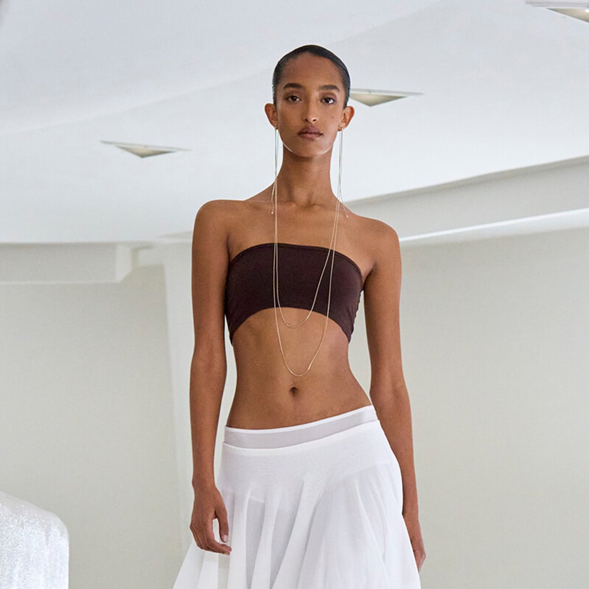 Model wearing a brown strapless bandeau top and white flowing skirt with long layered necklaces in a bright, minimalist setting.