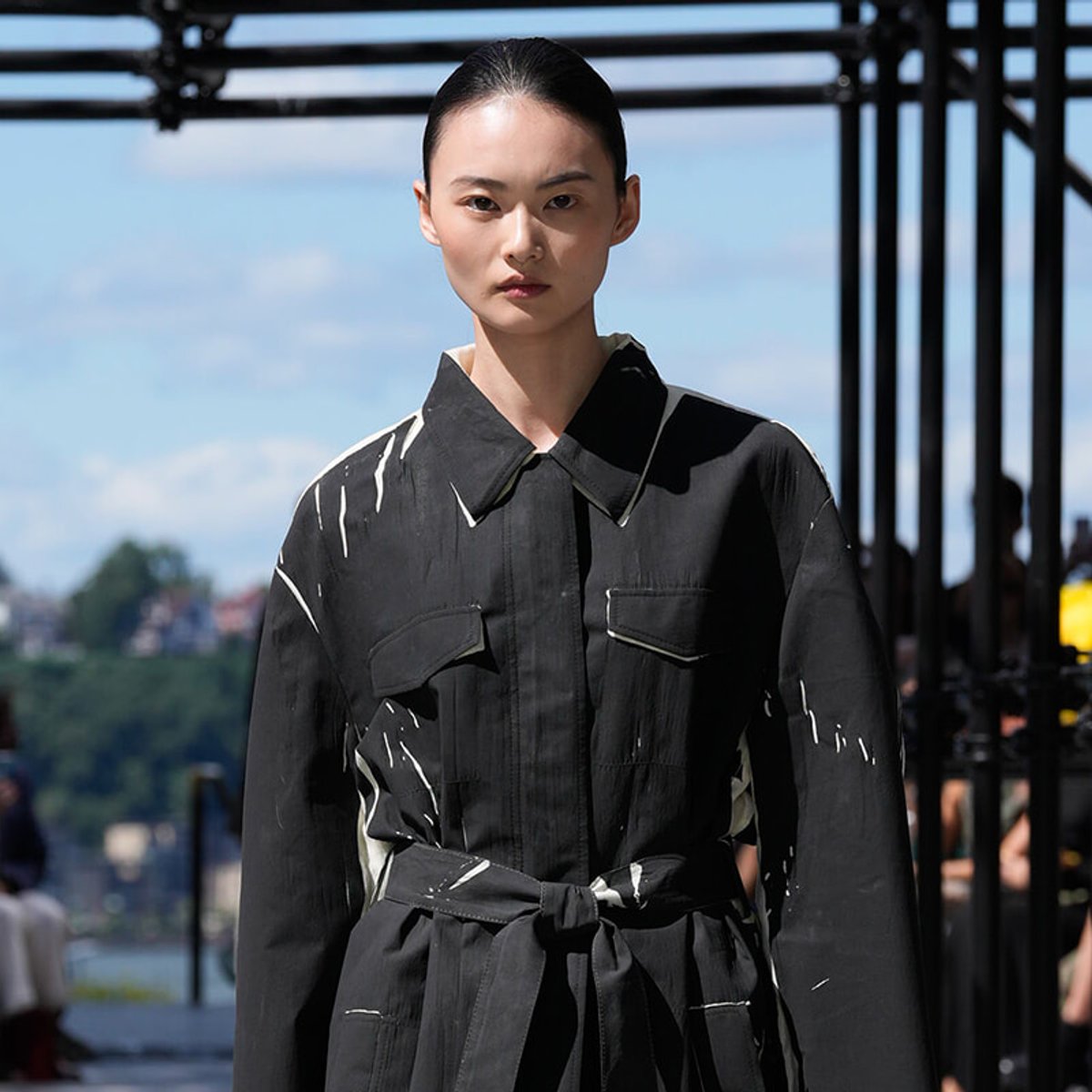 Model walking on runway wearing a black belted coat with white paint-splatter accents outdoors.