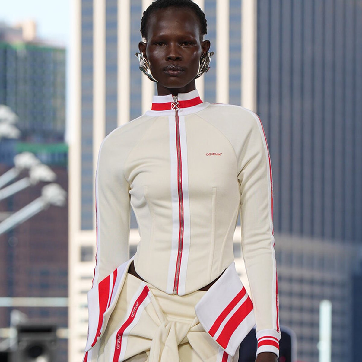 Model wearing a cream racing-inspired jumpsuit with red stripes and oversized collar on a city rooftop runway