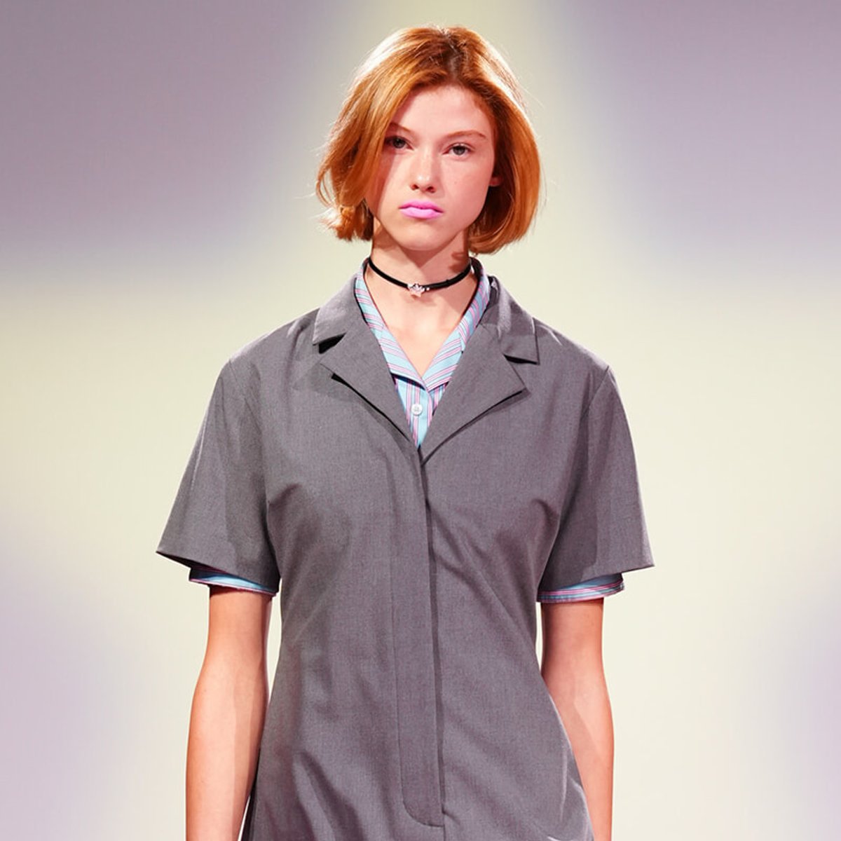 Model with short orange hair walking a fashion runway in a gray oversized coat over a striped shirt.