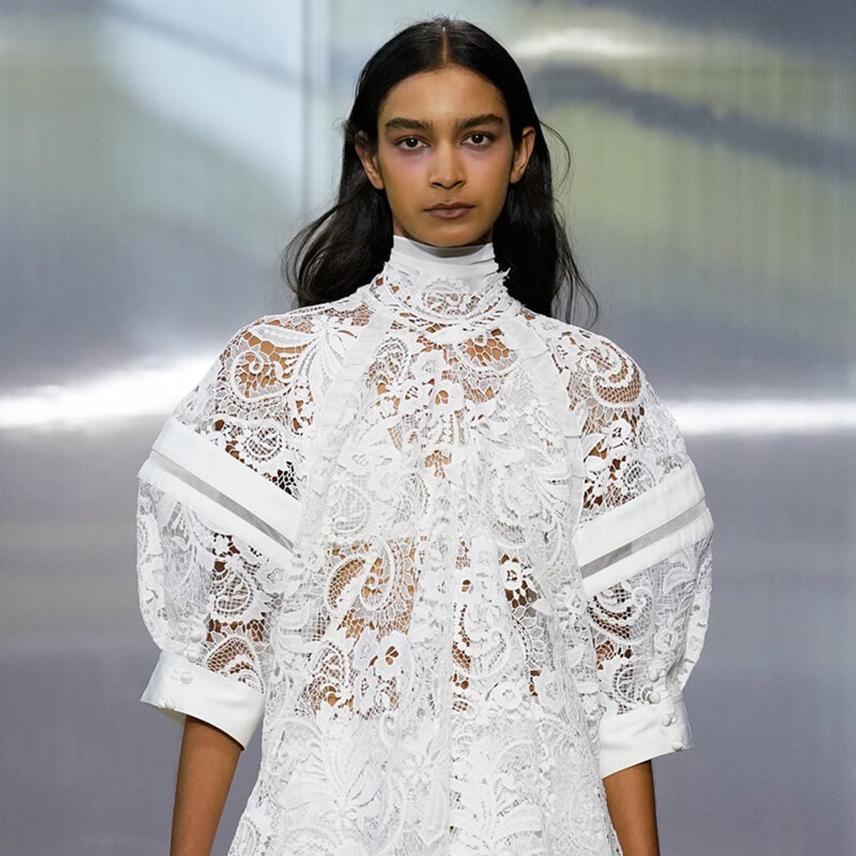 Female model wearing an intricate white lace high-neck dress on a runway.
