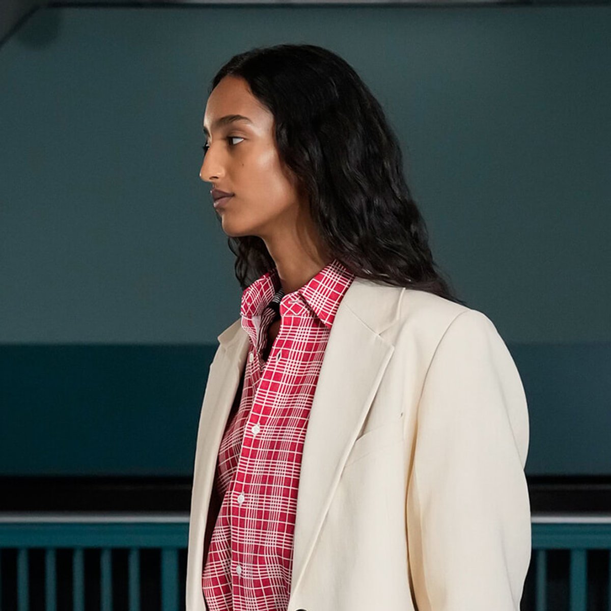 Woman in a cream blazer and red plaid shirt, seen in profile against a teal background.