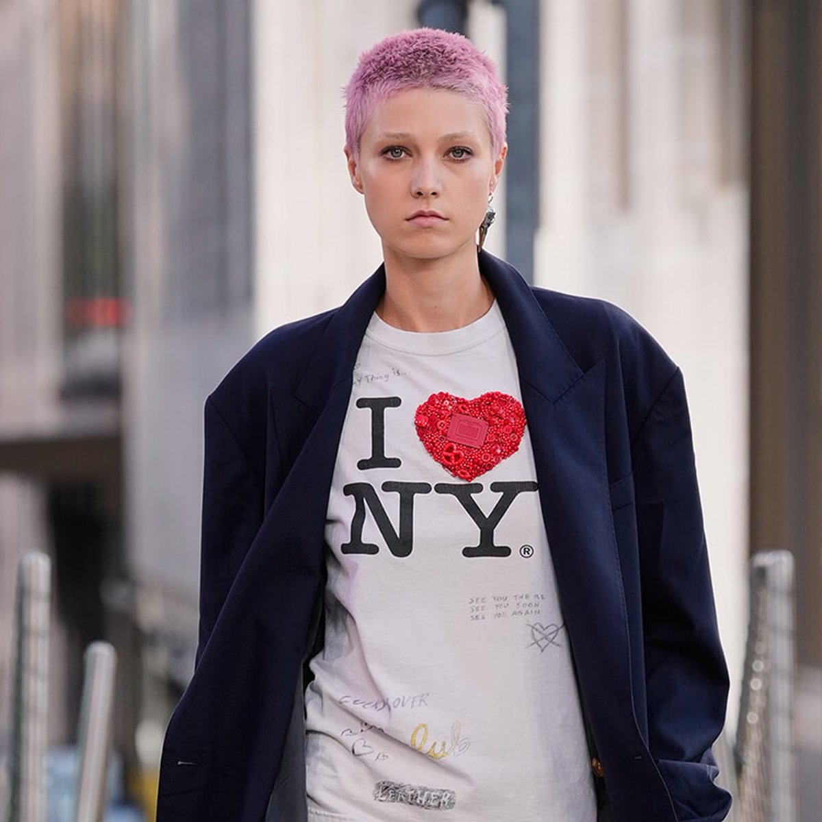 Person with short pink hair wearing a light gray 'I heart NYC' T-shirt and a dark blazer, walking down a city street.