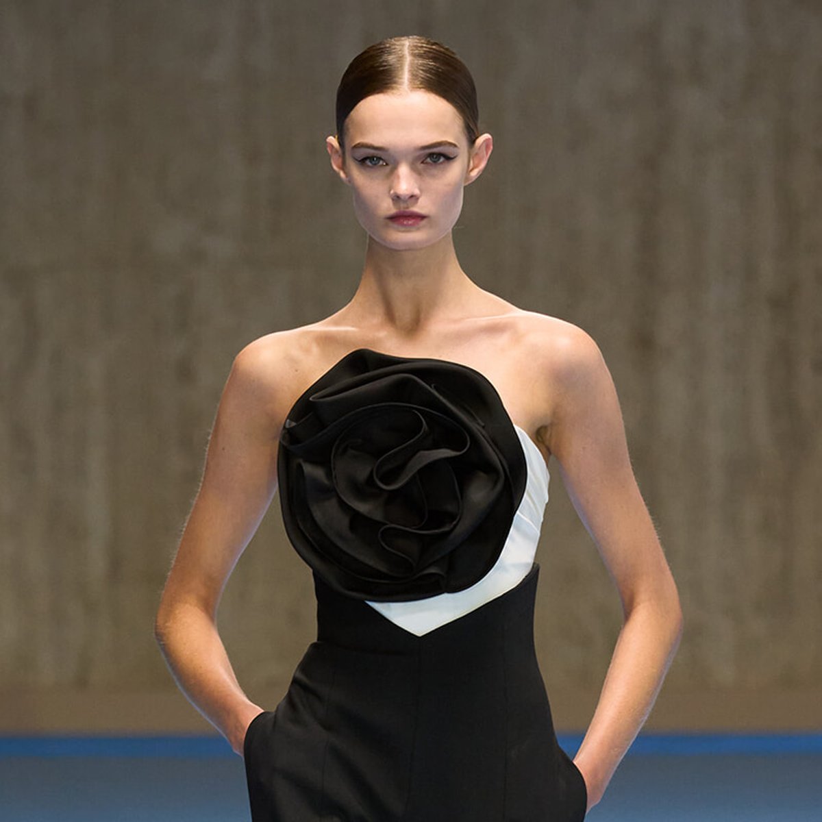 Model on a runway wearing a black strapless dress with a large black fabric rose on the bodice, hands in pockets.
