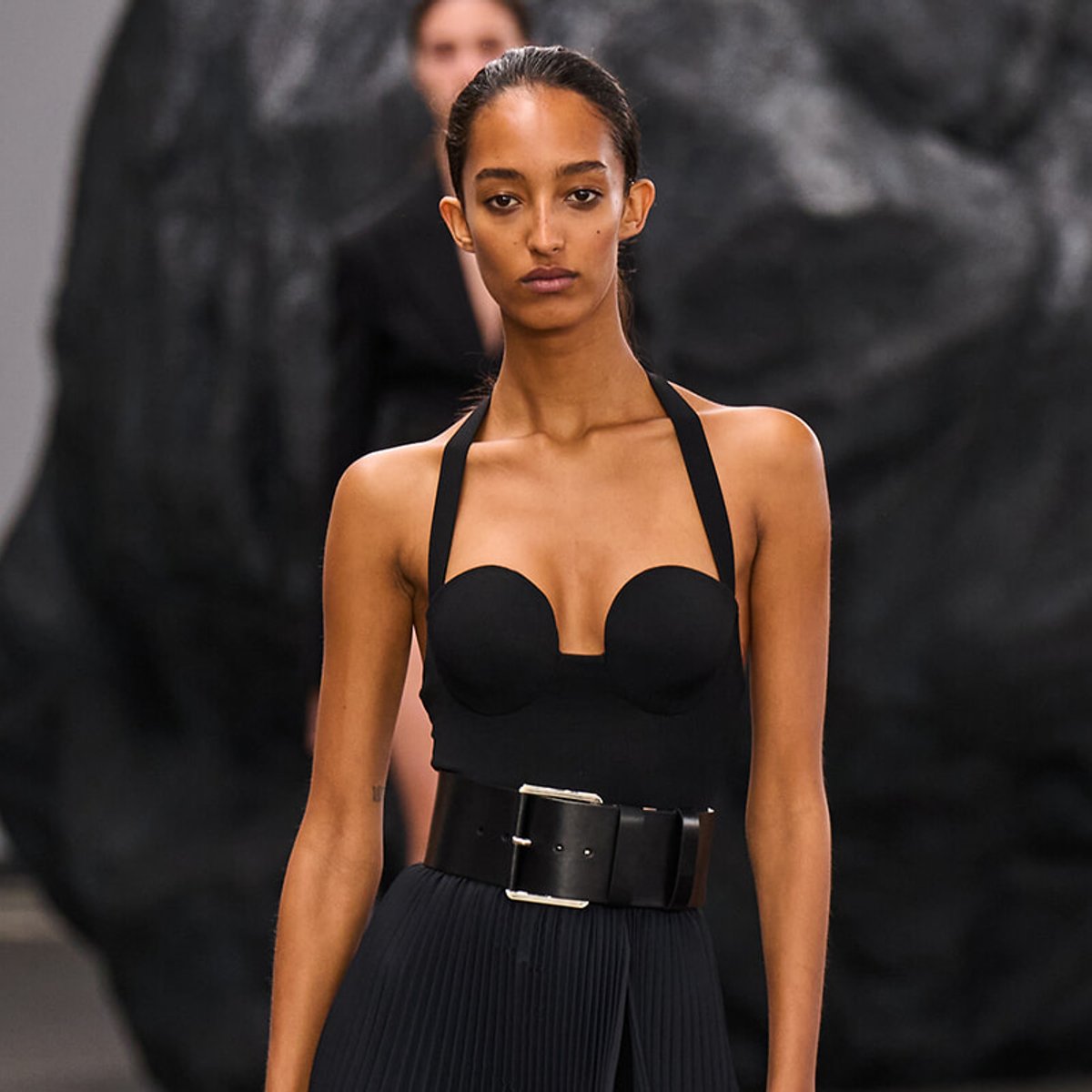 Model walking a fashion show runway in a black halter top with a wide belt and pleated skirt, against a dark backdrop.