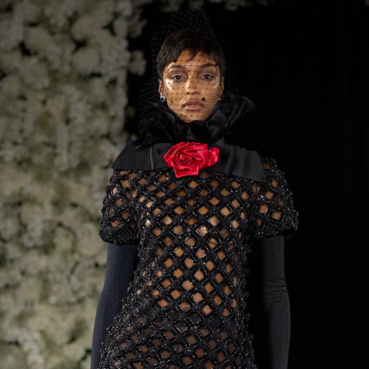 Model walking the runway in a black lattice dress with a red rose corsage and a black net veil.