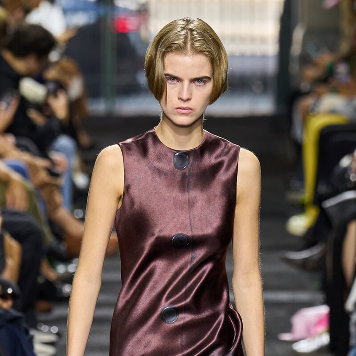 Model walking a runway in a burgundy satin sleeveless dress with large black buttons on the front.