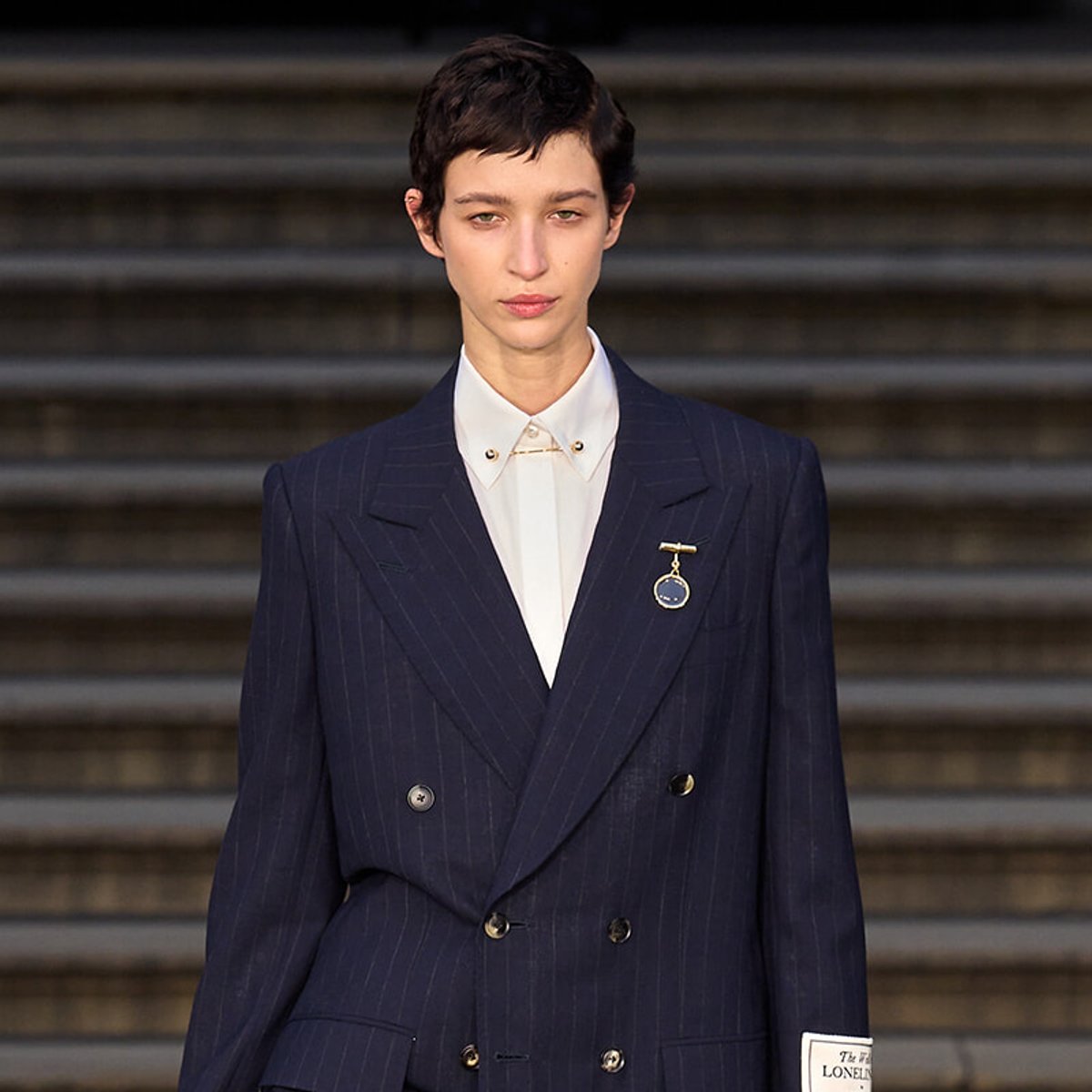 Model wearing a navy pinstripe double-breasted suit with a white shirt and lapel pin, standing on steps at a fashion show