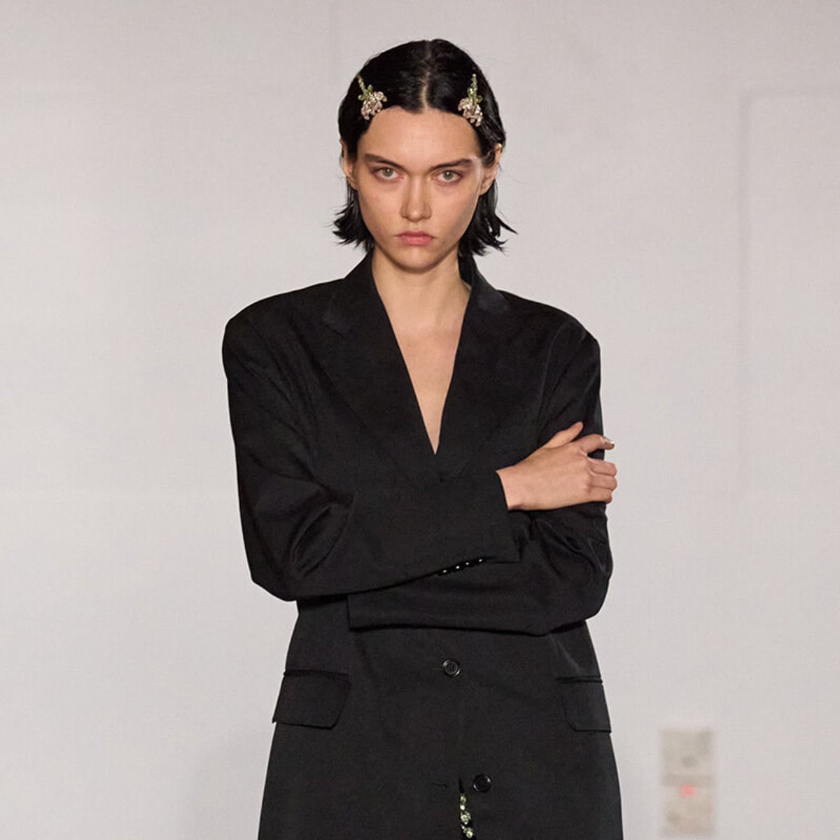 Female model on a runway wearing a black blazer with arms crossed and floral hairpins in her hair.