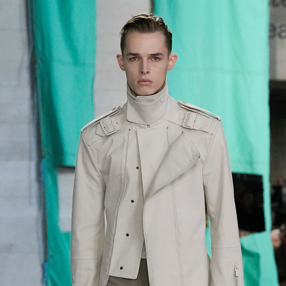 Male model wearing a light gray, high-collar avant-garde jacket with zippers and shoulder straps on a runway.