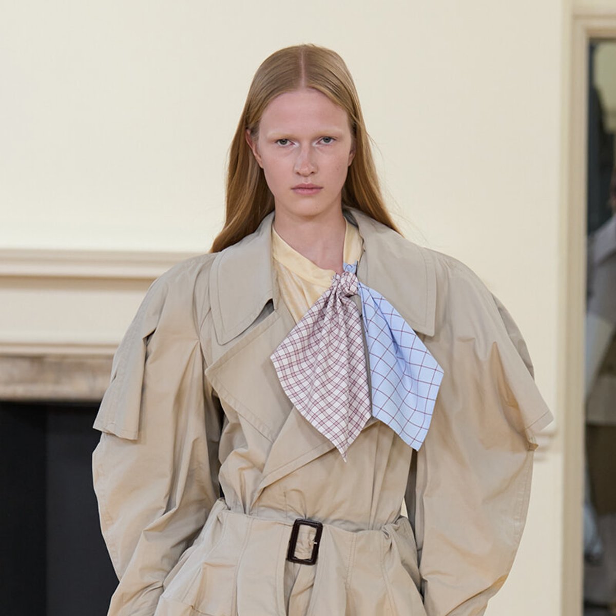 Model on a runway wearing a beige oversized trench coat with a belted waist and a two-toned checkered scarf tied at the neck.