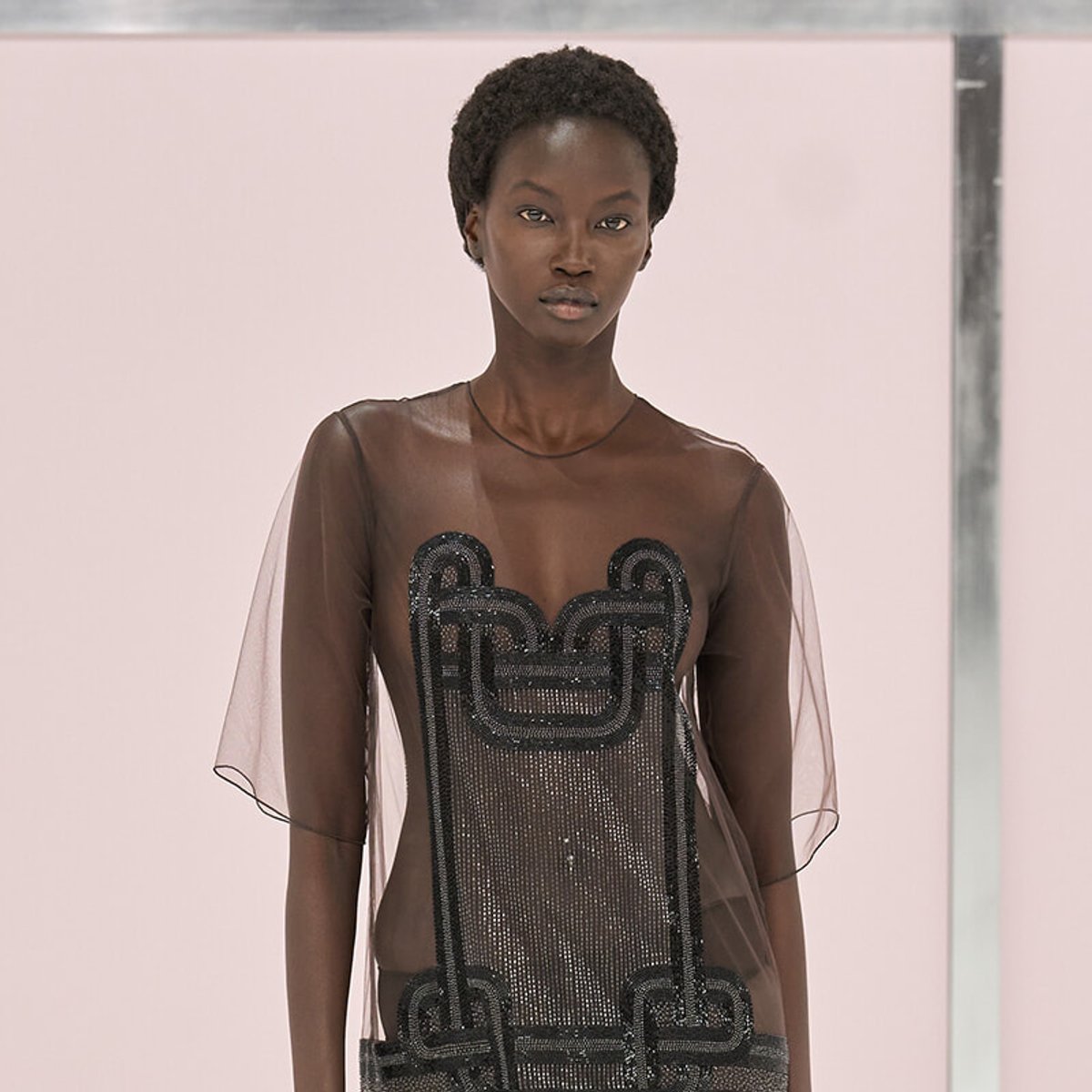 Runway model wearing a sheer brown mesh dress with black beaded geometric embroidery, standing against a pale backdrop.