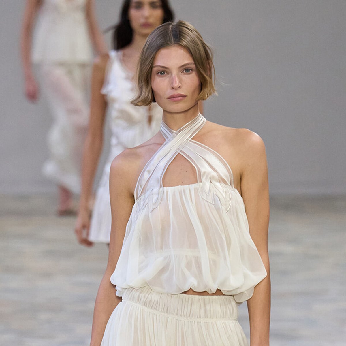 Model on a runway wearing a sheer white halter-top dress with crisscross straps and a gathered waist in a fashion show setting.