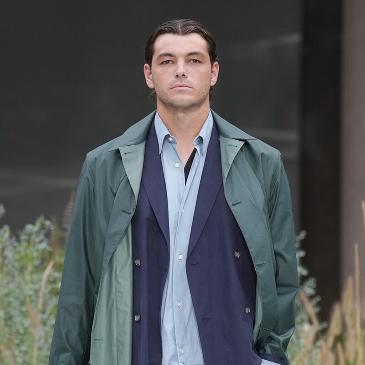 Man wearing a teal-green raincoat over a navy blazer and light blue shirt, standing outdoors with a neutral expression.