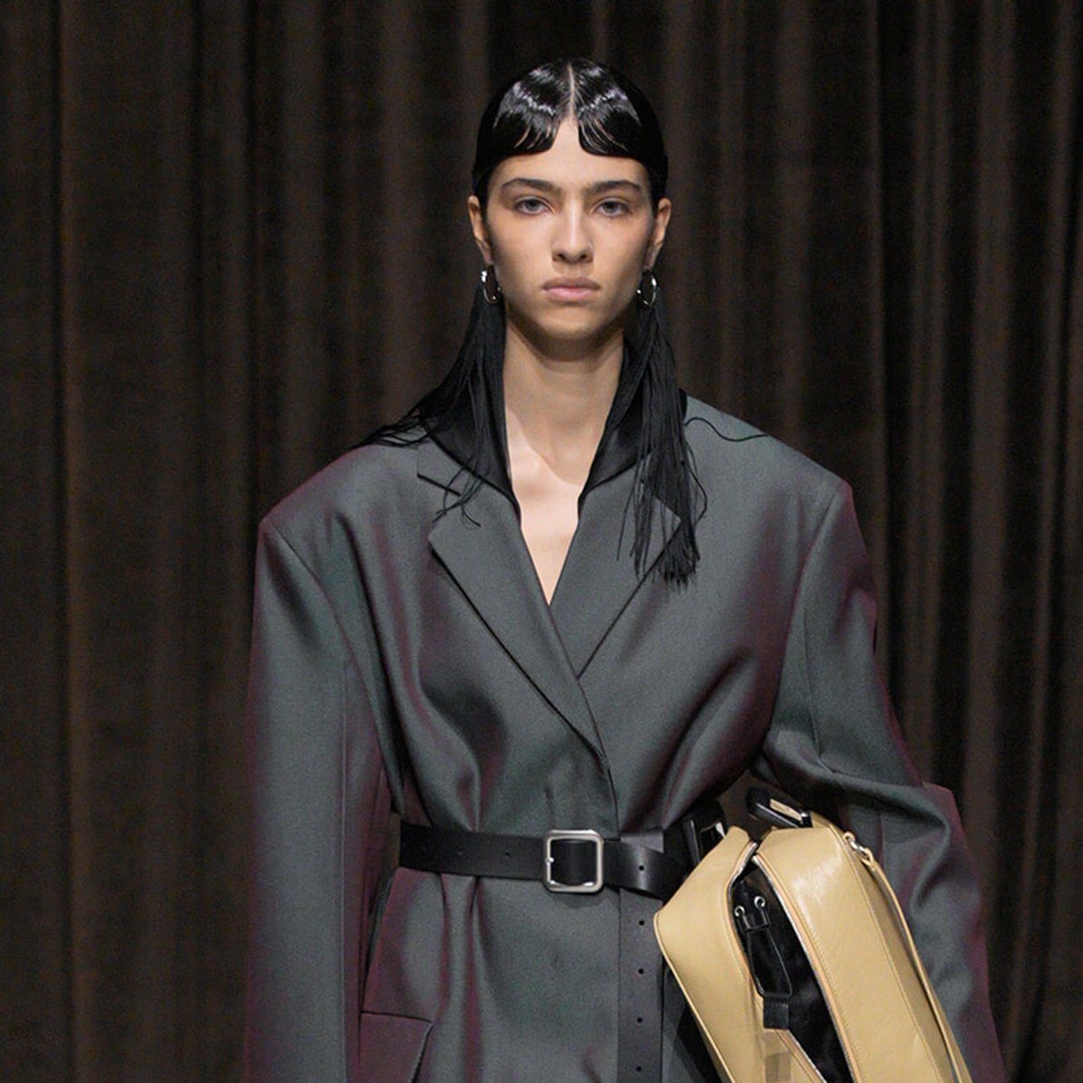 Model on a runway wearing an oversized gray blazer-style coat cinched with a black belt, holding a beige bag, with slicked-back black hair and a neutral expression against a dark backdrop.