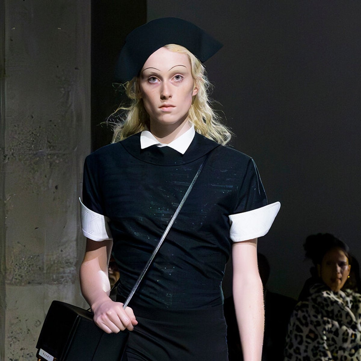 Runway model in a black dress with white collar and cuffs, black hat, and a crossbody bag, walking the catwalk.