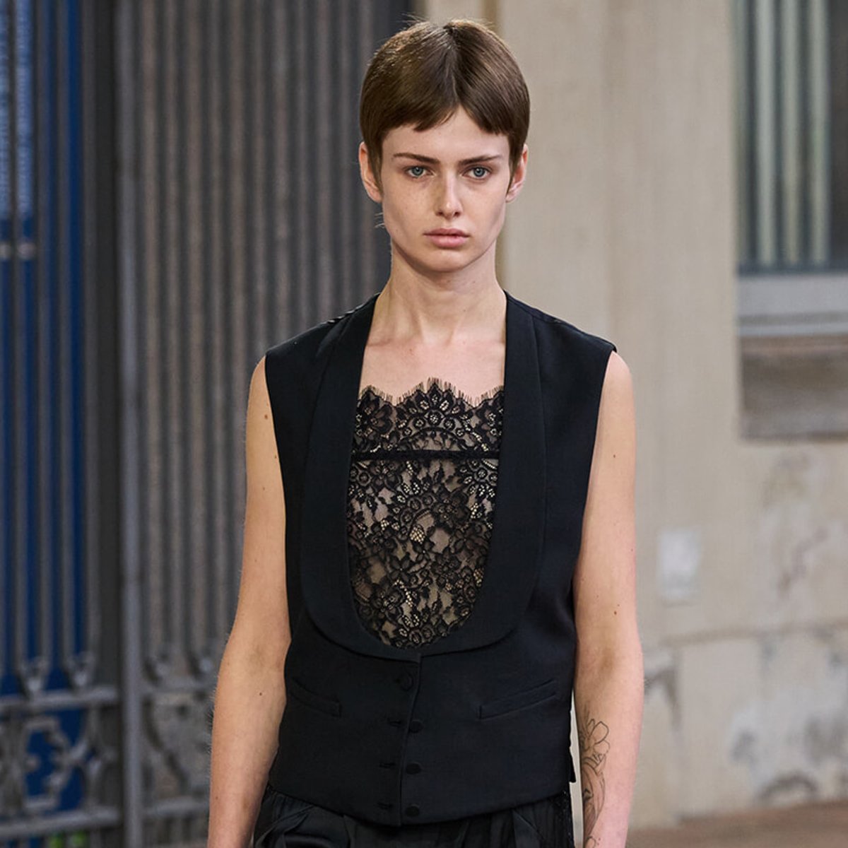 Model wearing a black lace top with a matching black vest, standing in front of an urban backdrop with neutral expression.