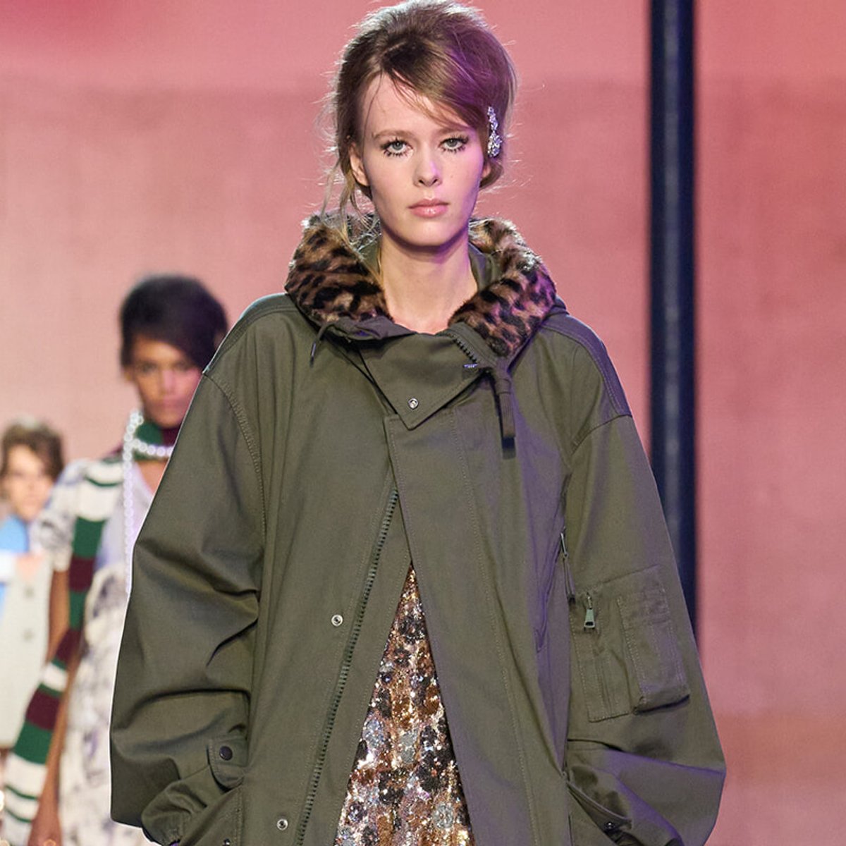 Model walks a fashion runway in an oversized olive-green parka with a leopard-print fur collar, blurred background models behind.