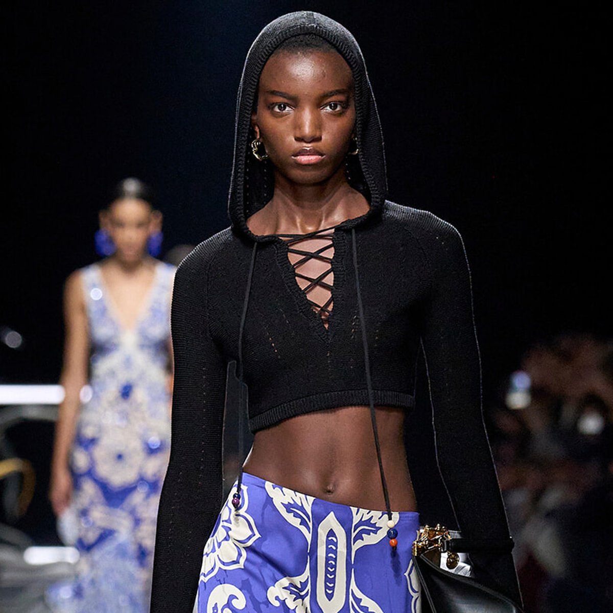 Runway model wearing a black knit cropped hoodie with a lace-up front, paired with a blue and white patterned skirt.