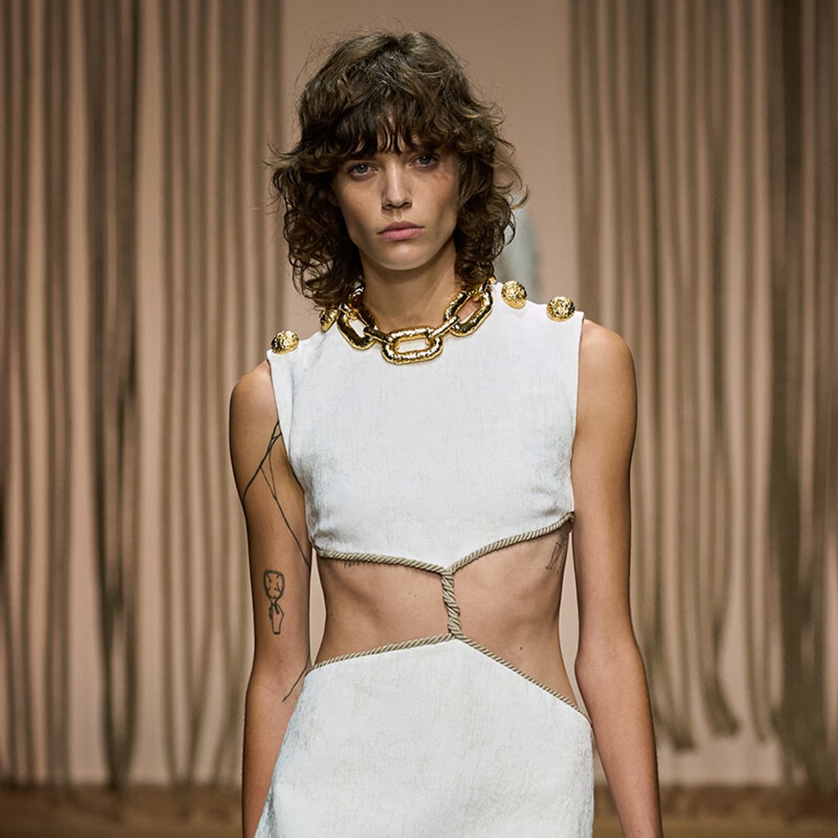 Female model on a runway wearing a sleeveless white dress with cutouts and rope waist detailing, accessorized with a chunky gold chain necklace, against a neutral backdrop.