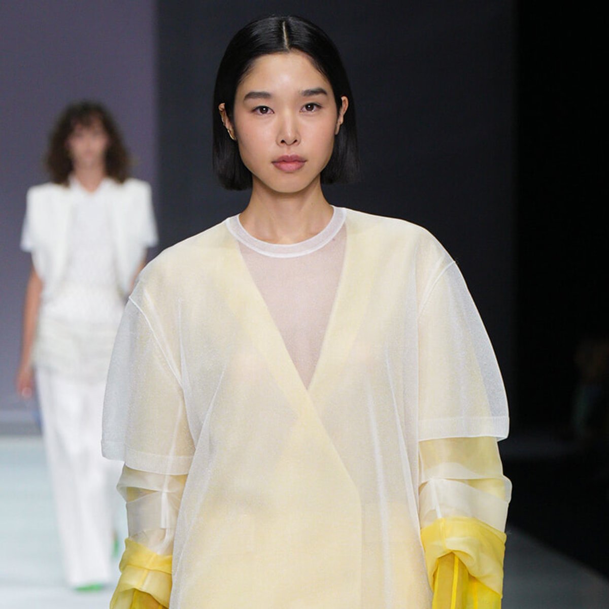 Asian female model walks the runway in a sheer pale yellow-white top; background model in white walks behind.