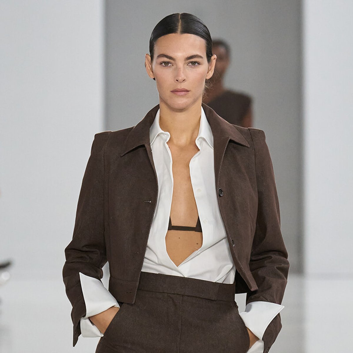Female model walking a runway in a brown suit with a white shirt open at the chest, revealing a black bralette beneath.