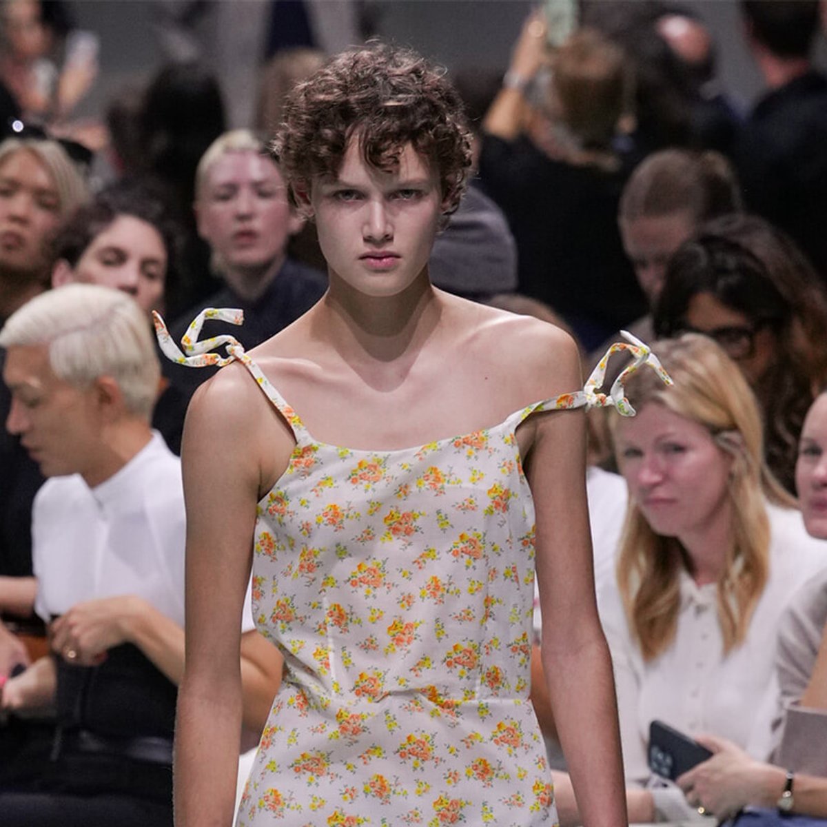 Model walking a runway in a white floral sundress with tying straps, audience blurred in the background.