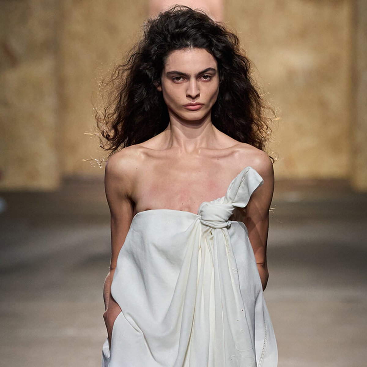 Model walking runway in a white, one-shoulder dress tied in a knot at the bust, confident expression, stone backdrop behind her