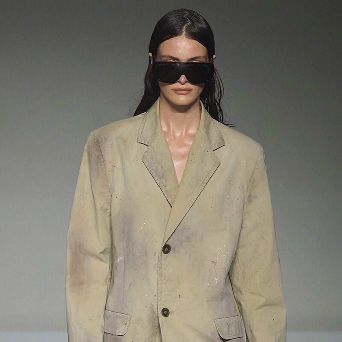 Model on a runway wearing an oversized beige blazer with paint splatters and large dark sunglasses.