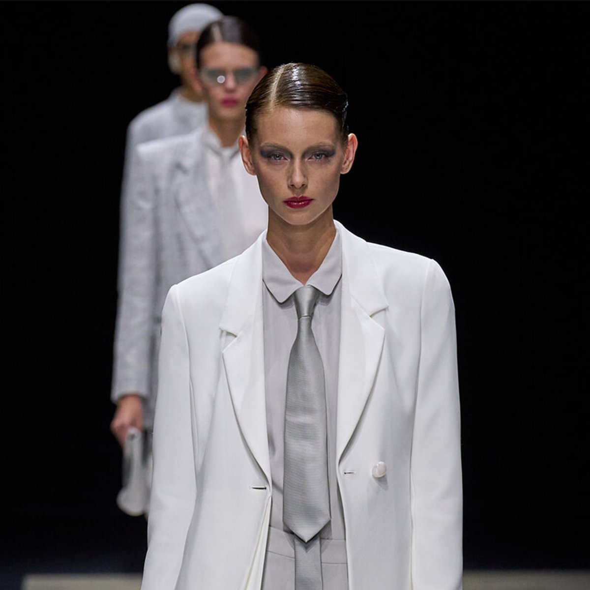 Model on a runway wearing a white blazer and matching suit with a gray tie, slicked hair, and bold makeup.