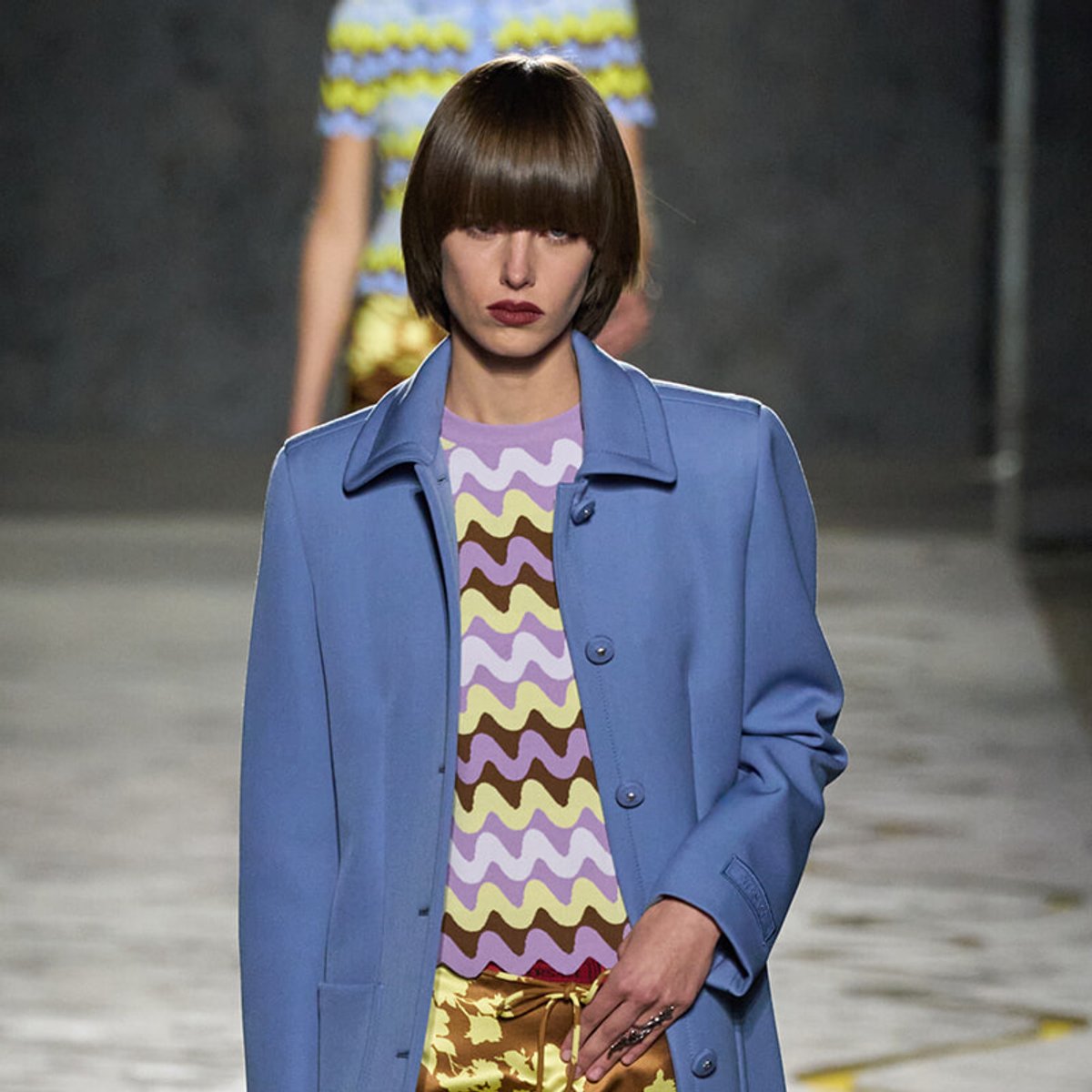 Model walking runway in a light blue coat over a purple, yellow, and brown zigzag top; blurred background.