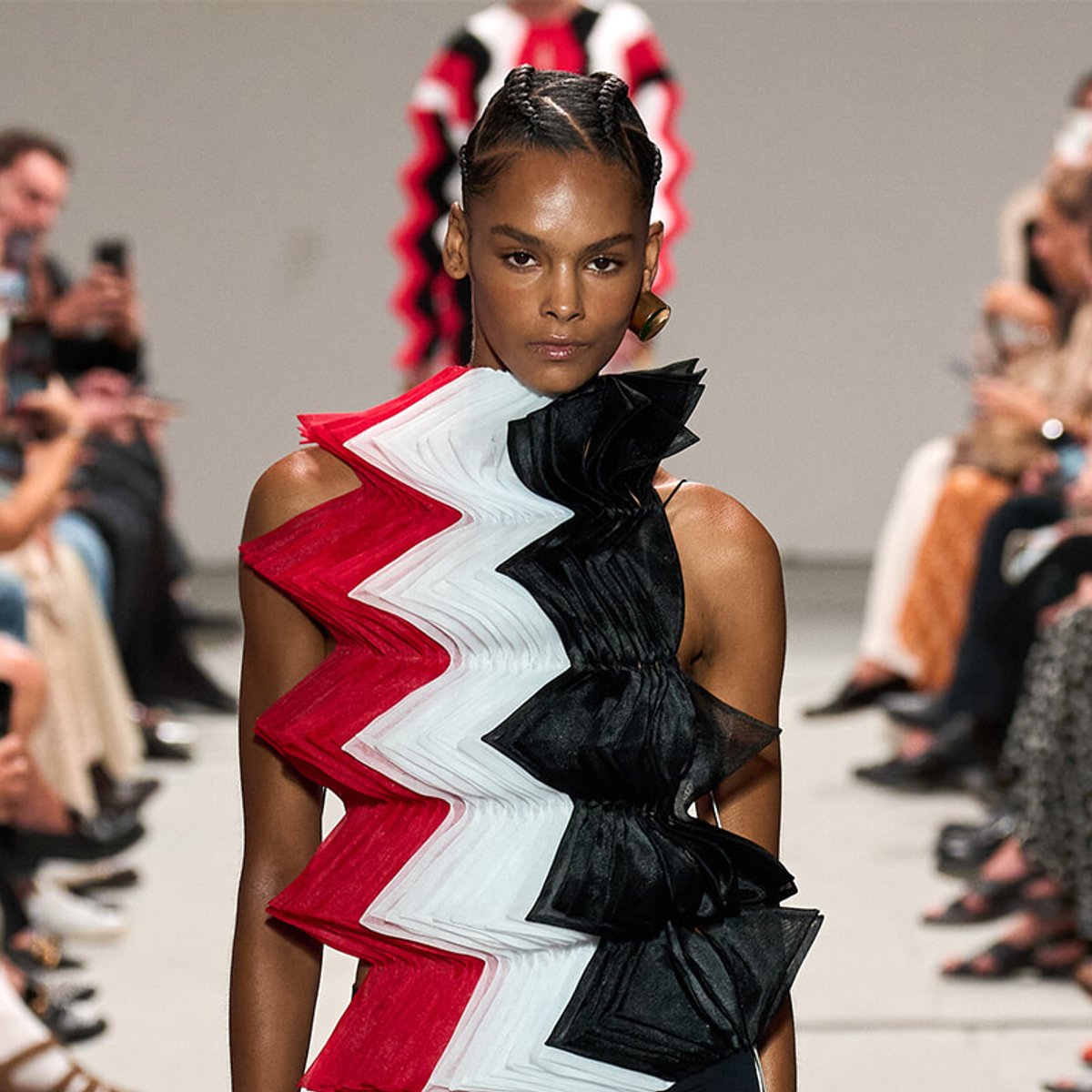 Model walking a runway in a bold red, white, and black zigzag ruffle dress; audience blurred in background.