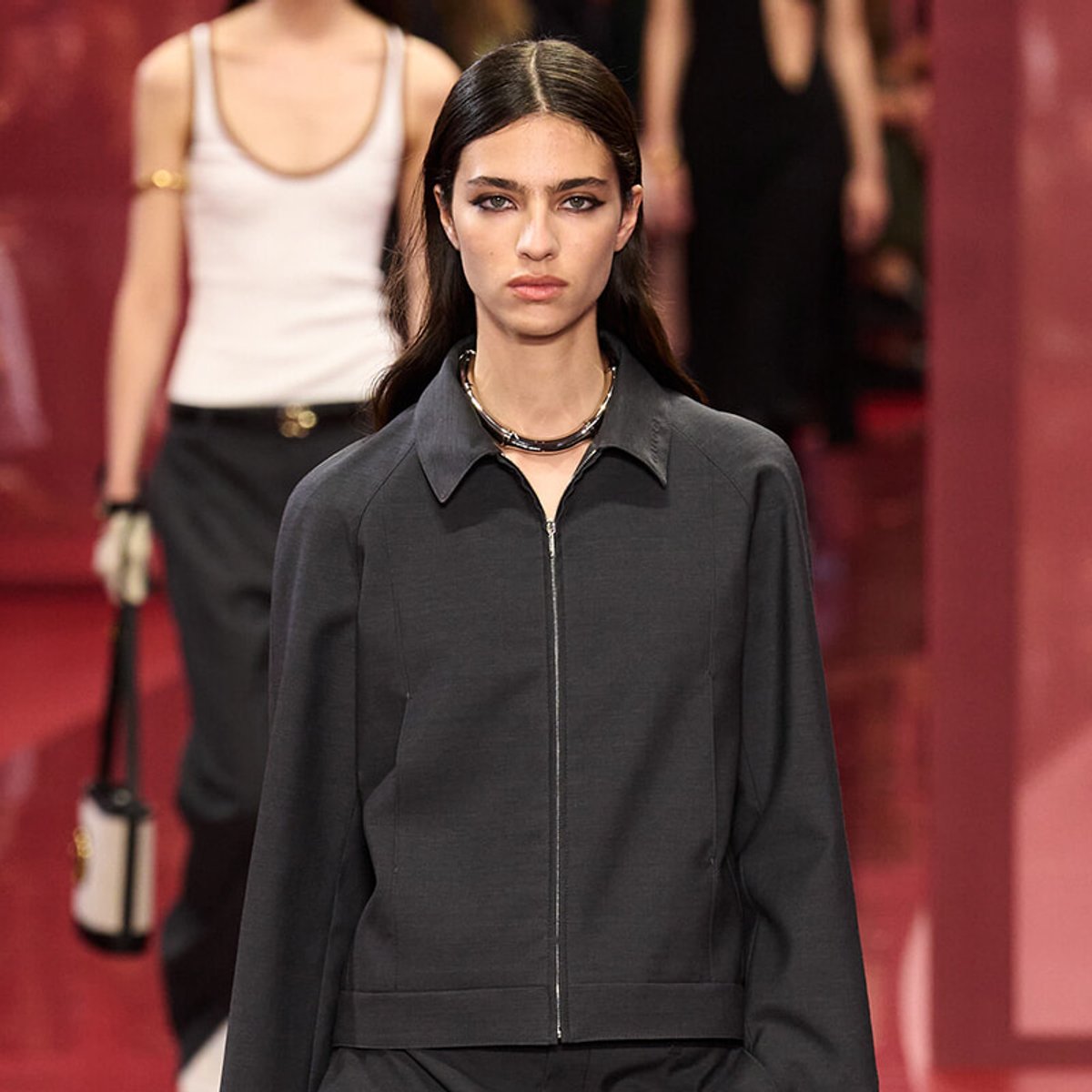 Model walking a fashion runway in a dark zip-front jacket and layered necklace.