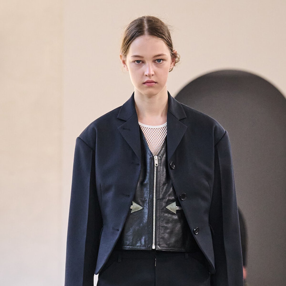 Female model on a runway wearing a navy blazer over a black leather vest and mesh top, with a neutral expression.