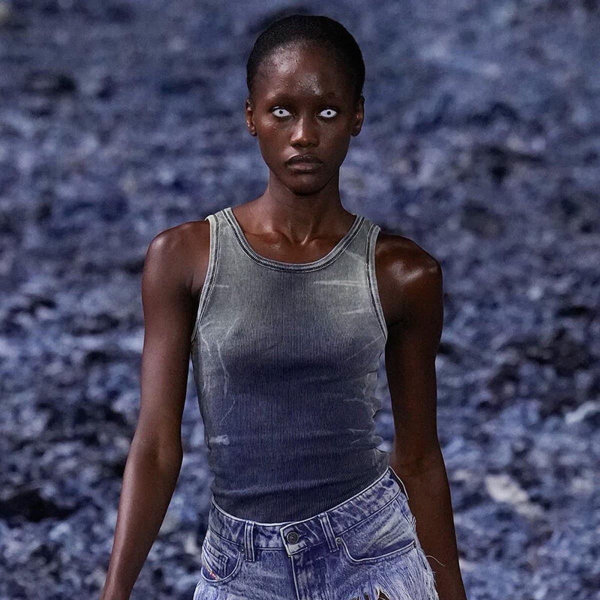 Model with dark skin and white-glazed eyes wearing a gray sleeveless top and denim jeans, standing against a blue rocky background ahead.