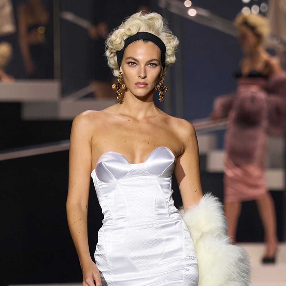 Model walking the runway in a white strapless satin dress with a corset-style bodice, black headband, and large gold earrings, holding a white fur stole.