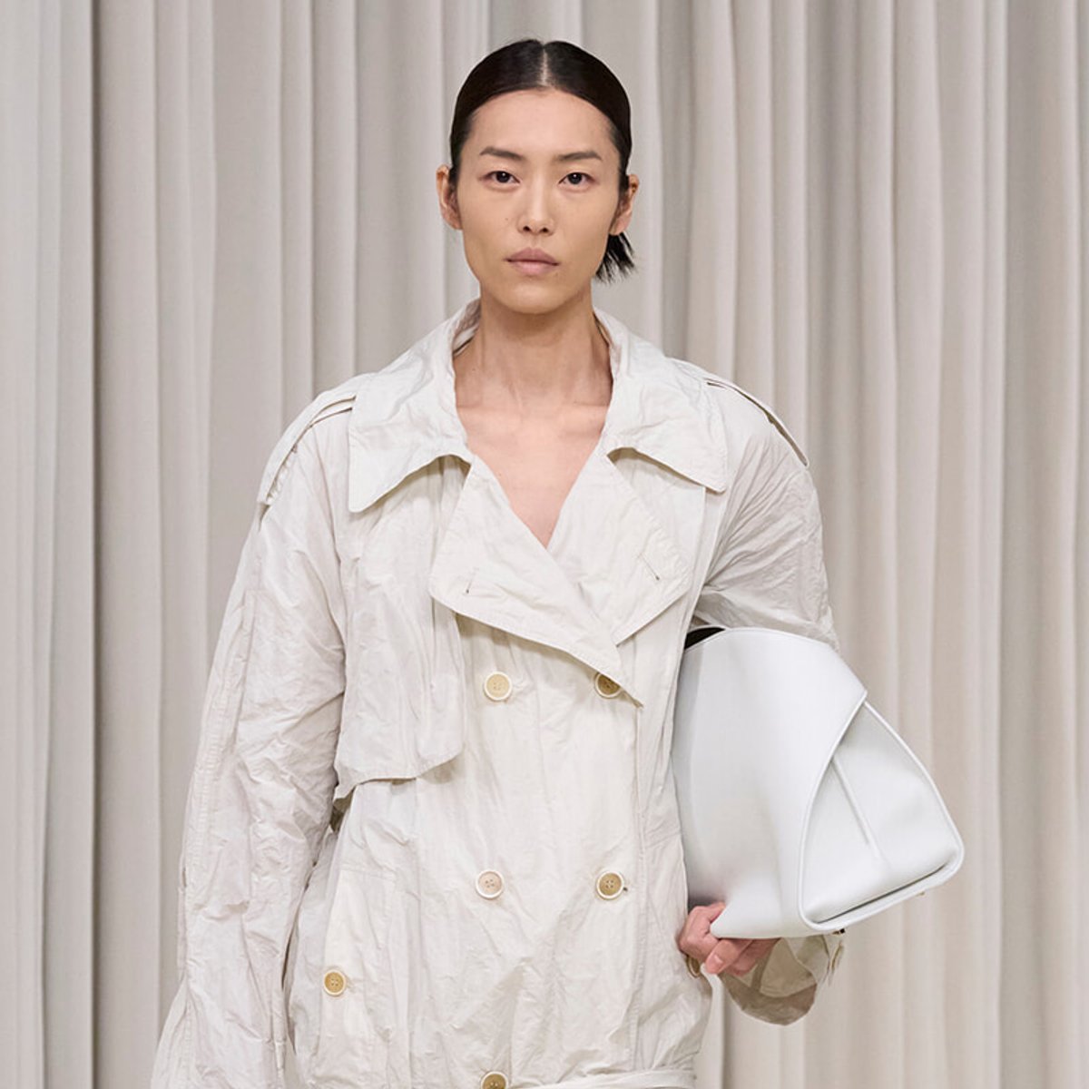 Model wearing a crinkled beige trench coat, holding a white handbag against a neutral curtain backdrop.d