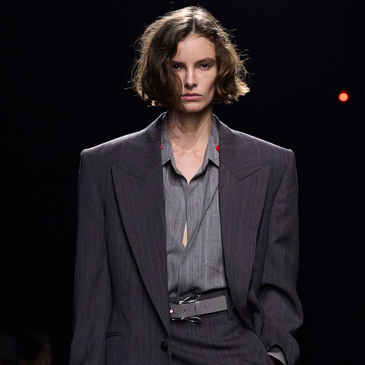 Model on runway wearing a dark pinstripe suit over a gray shirt, with a wide belt, looking straight ahead.