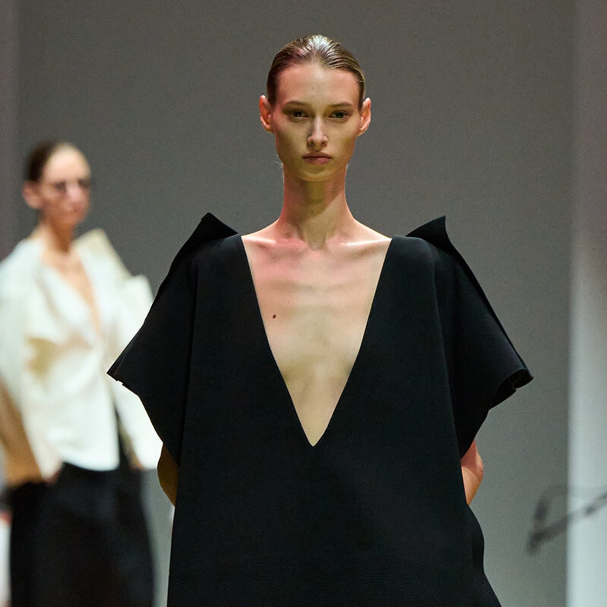 Model on a runway wearing a black deep-V dress with sculpted, angular shoulders.