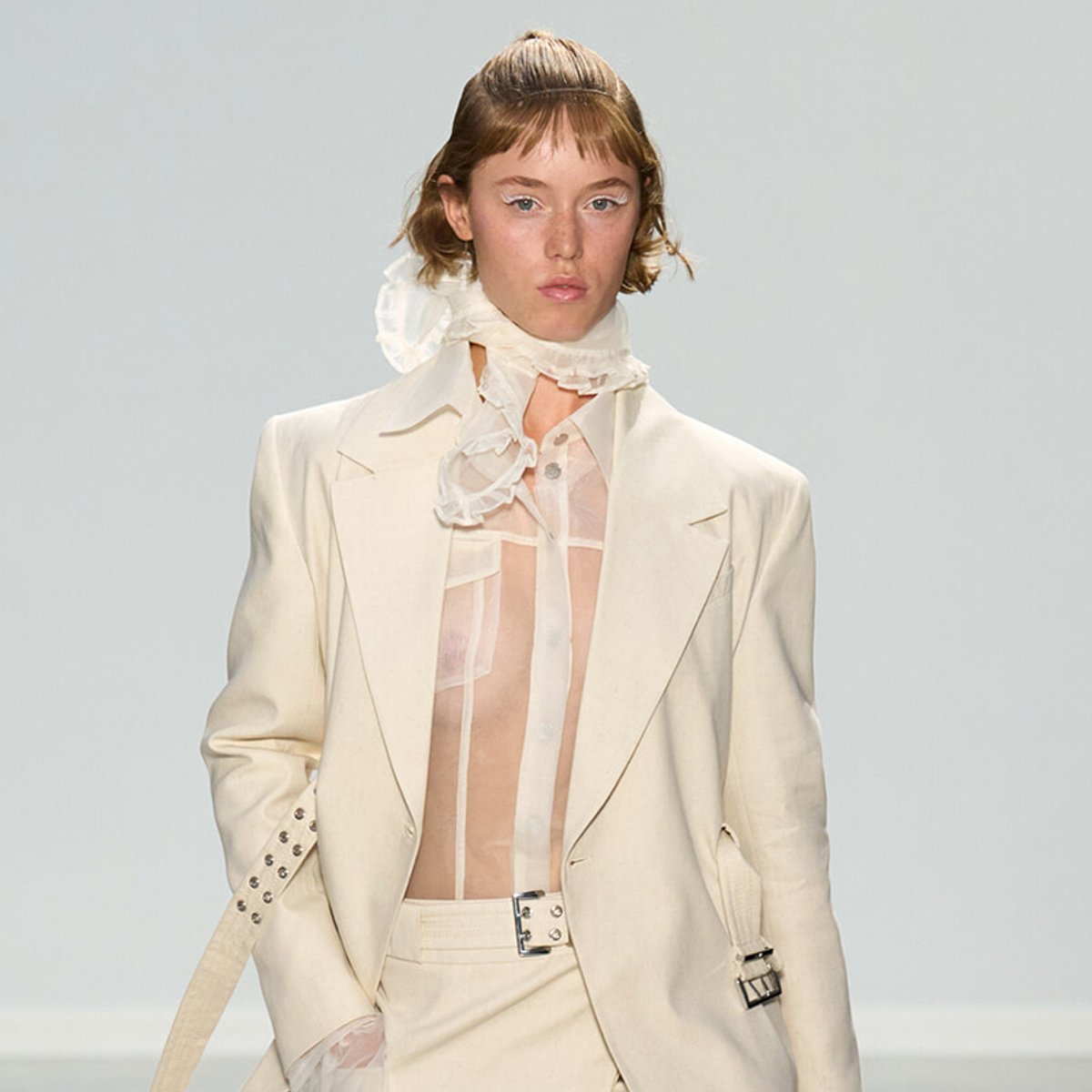 Female model walking a runway in a cream suit with sheer blouse and ruffled scarf detail at the neck.
