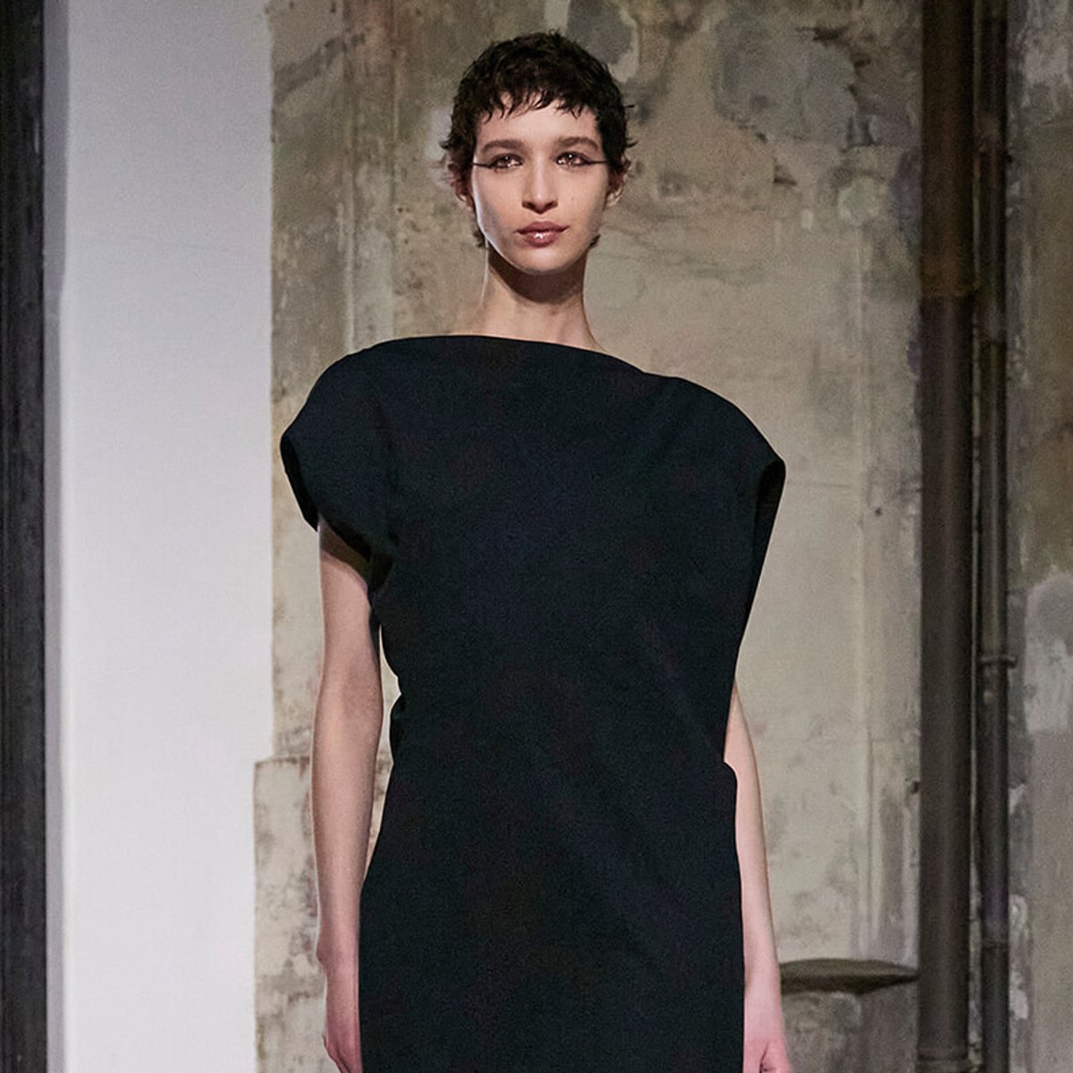 Model wearing a minimalist black sheath dress on a runway with an industrial, textured backdrop.