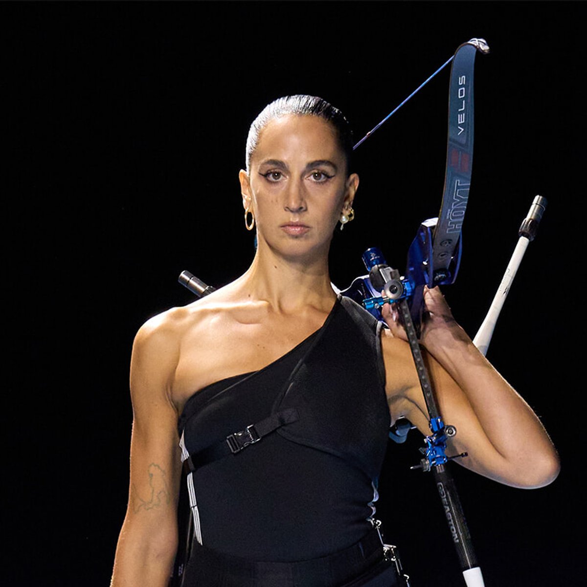 Female archer in a one-shoulder black outfit, holding a modern compound bow over her shoulder with a focused expression.