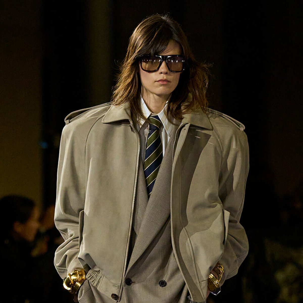 Model wearing a beige trench coat, white shirt, striped tie and dark sunglasses on a runway, with gold bracelets visible on wrists.