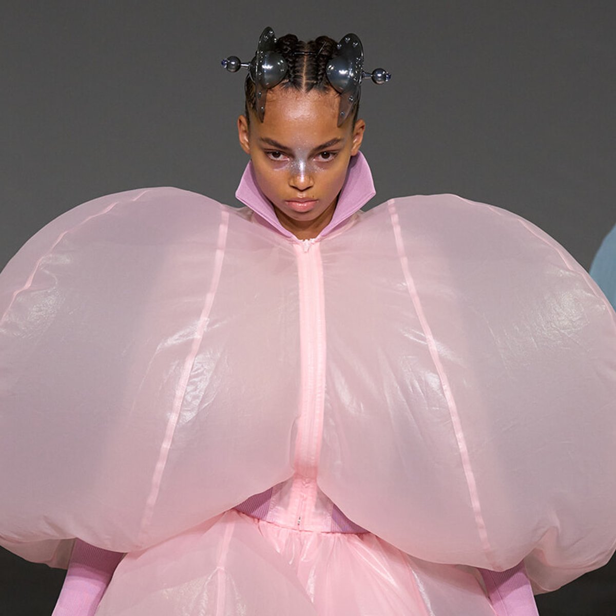 Model on a runway wearing a pastel pink, oversized voluminous outfit with giant sleeve-like shapes and a futuristic headpiece, looking forward.