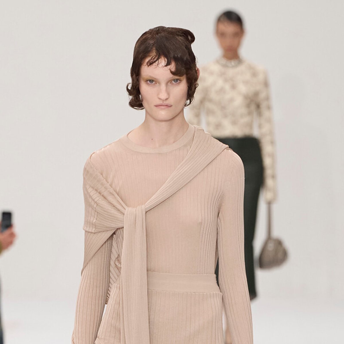 Runway model in a beige ribbed knit dress with a draped scarf over the shoulder; a second model is blurred in the background.