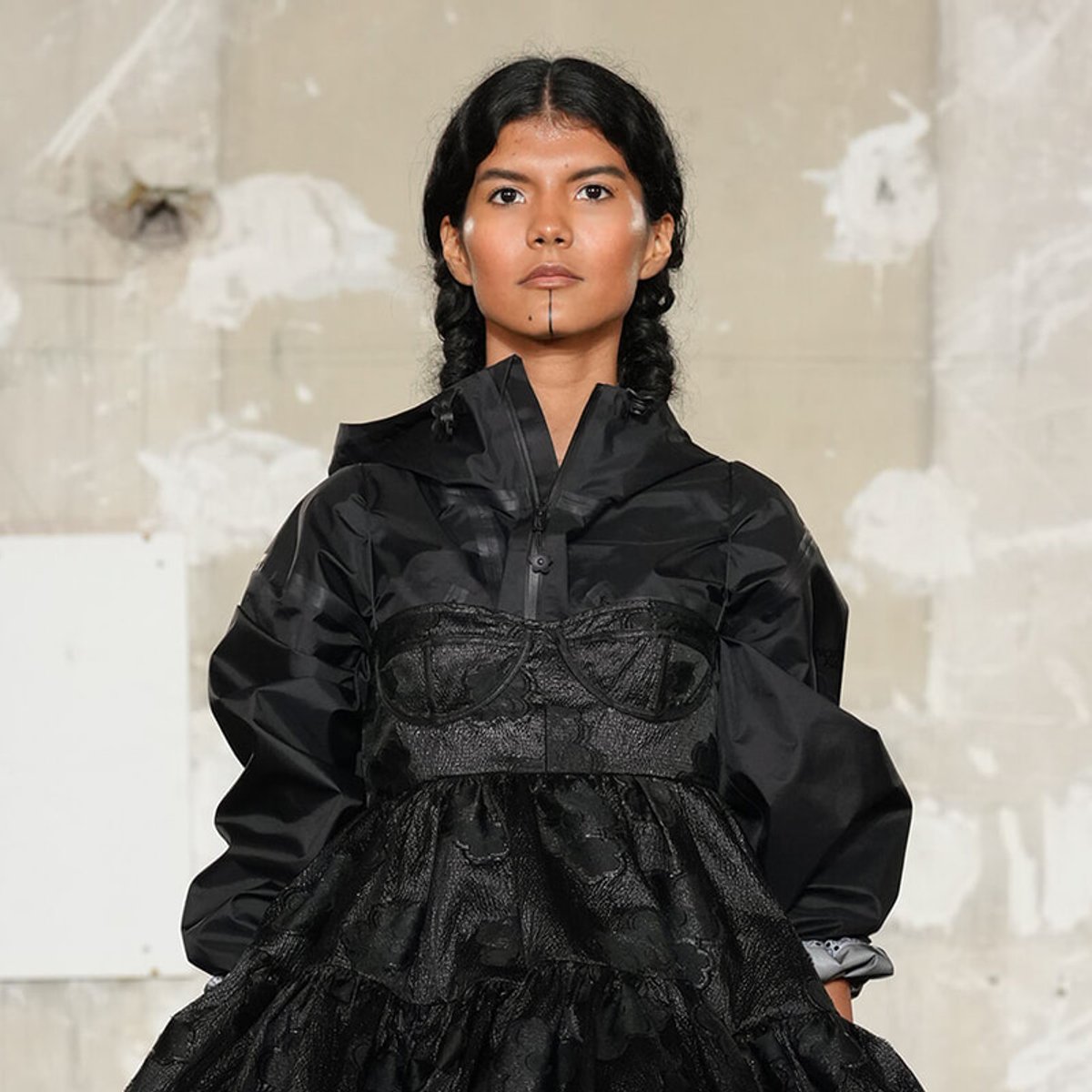 Model in a black layered dress with a lace corset over an oversized jacket on a runway, looking forward with a neutral expression.
